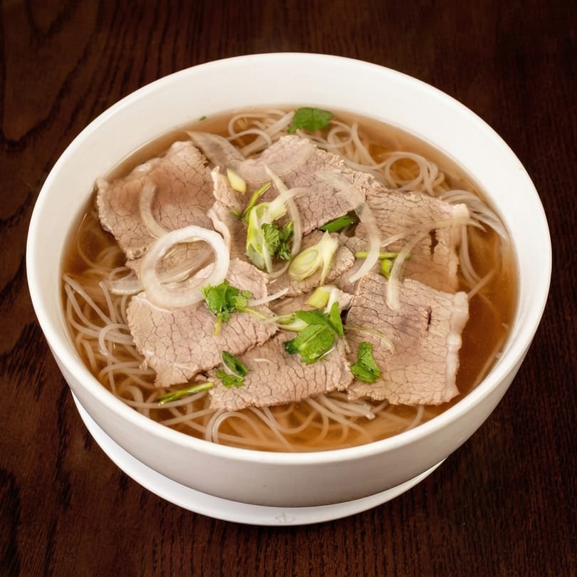 A white bowl of pho with thinly sliced beef, rice noodles, broth, onions, and cilantro.