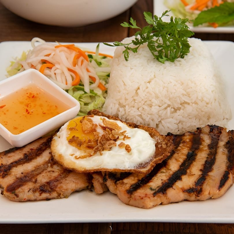 Grilled pork, fried egg, white rice, pickled vegetables, and dipping sauce on a white plate.
