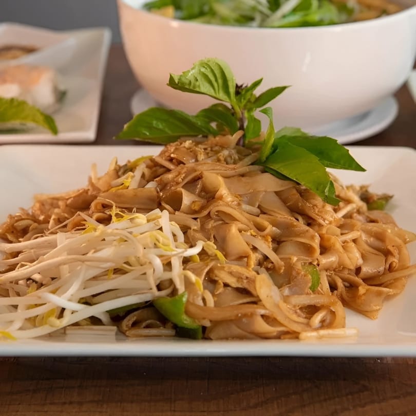 Close-up of a plate of Pad See Ew noodles with bean sprouts and basil, with other dishes blurred in the background.