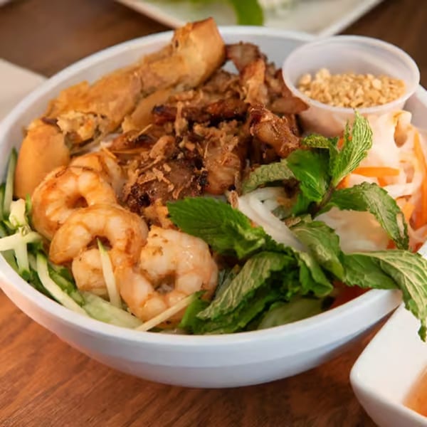 A white bowl filled with Vietnamese vermicelli noodles, grilled pork, shrimp, egg rolls, and fresh herbs.